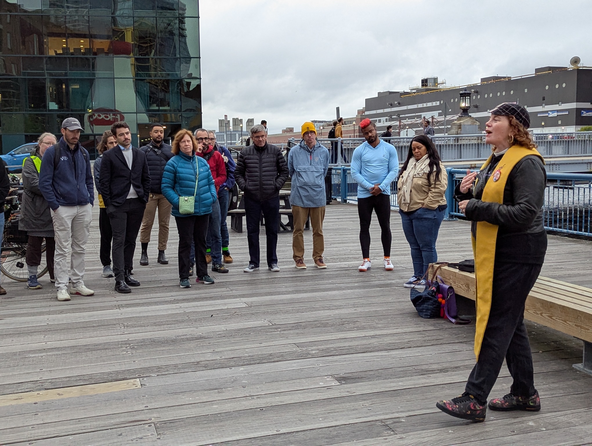 A woman wearing a yellow clerical stole over a leather jacket speaks in front of a small crowd of people on a city plaza.