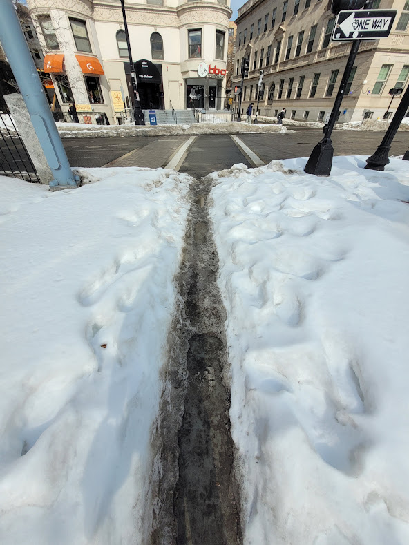 A busy crosswalk at Kenmore Street and Commonwealth Avenue covered in deep slush and snow, with a narrow, muddy, human-made path.